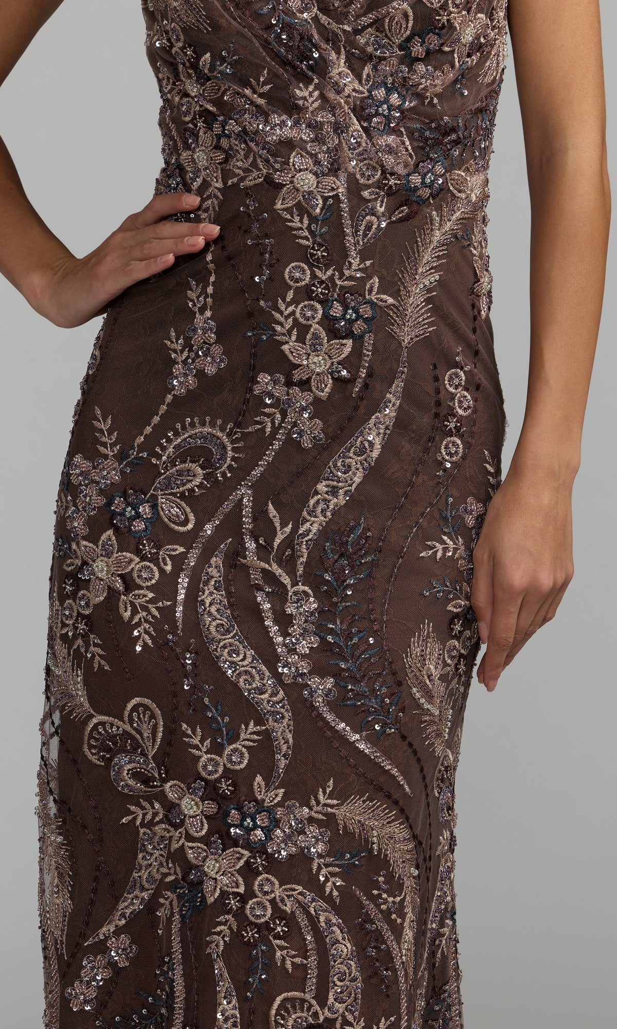 A person wearing the Mac Duggal 68914 long formal dress, a sleeveless brown gown adorned with intricate floral and sequin embroidery, stands with one hand on their hip.